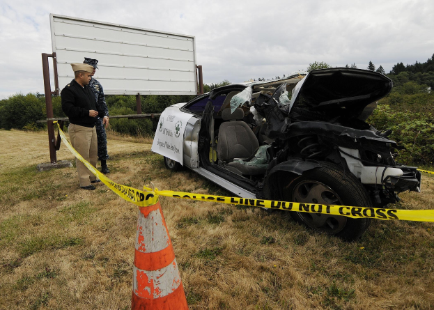 Ein Polizeibeamter in Uniform steht neben einem schwer beschädigten Auto am Straßenrand mit eingedöllter Haube und zersplitterten Fenstern, während in der Nähe ein Verkehrskegel und ein Absperrband an einem Pfosten gegen den Hintergrund von Bäumen und einem bewölktem Himmel zu sehen sind.