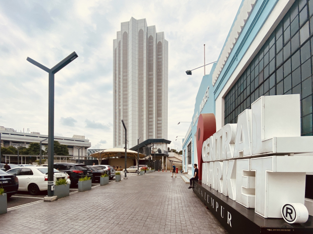Ein belebter zentraler Markt in Kuala Lumpur, Malaysia, mit hohen Gebäuden, Fahrzeugen, Fußgängern, Straßenmöbeln, Beschilderungen, Bäumen und einem bewölkten Himmel.