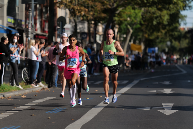 Gruppe von Menschen, die bei einem Stadtmarathon auf einer Straße mit Zuschauern laufen, unscharfer Hintergrund mit Bäumen, Gebäuden und einem Fahrrad.
