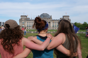 Drei Frauen in verschiedenen farbigen Kleidern, eine trägt eine Mütze, stehen vor dem Reichstaggebäude in Berlin, Deutschland, mit Menschen, Flaggen, Bäumen, Wolken und einem bewölkten Himmel im Hintergrund.