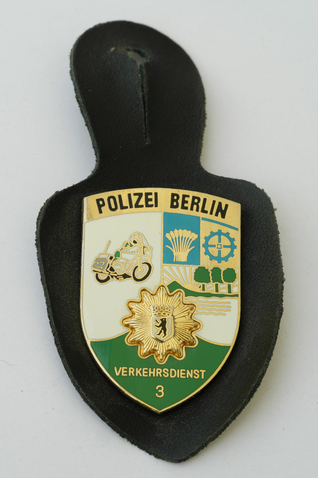 Ein Polizeistern mit der Aufschrift "Polizei Berlin", der ein blaues Schild mit einem weißen Stern in der Mitte zeigt, umgeben von einem weißen Rand, auf einer Oberfläche.
