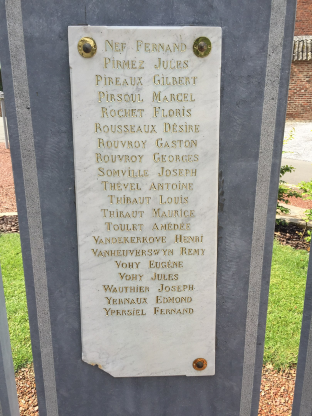 Steinbank mit einer Gedenktafel für Kriegsopfer, vor einem Backsteingebäude mit Bäumen und Gras.