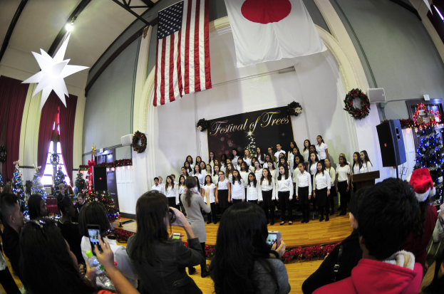 Gruppe von Menschen auf einer Bühne mit Weihnachtsdekoration, einem Rednerpult, Lautsprechern, einer Texttafel und einer Menge darunter mit Kameras und Handys.