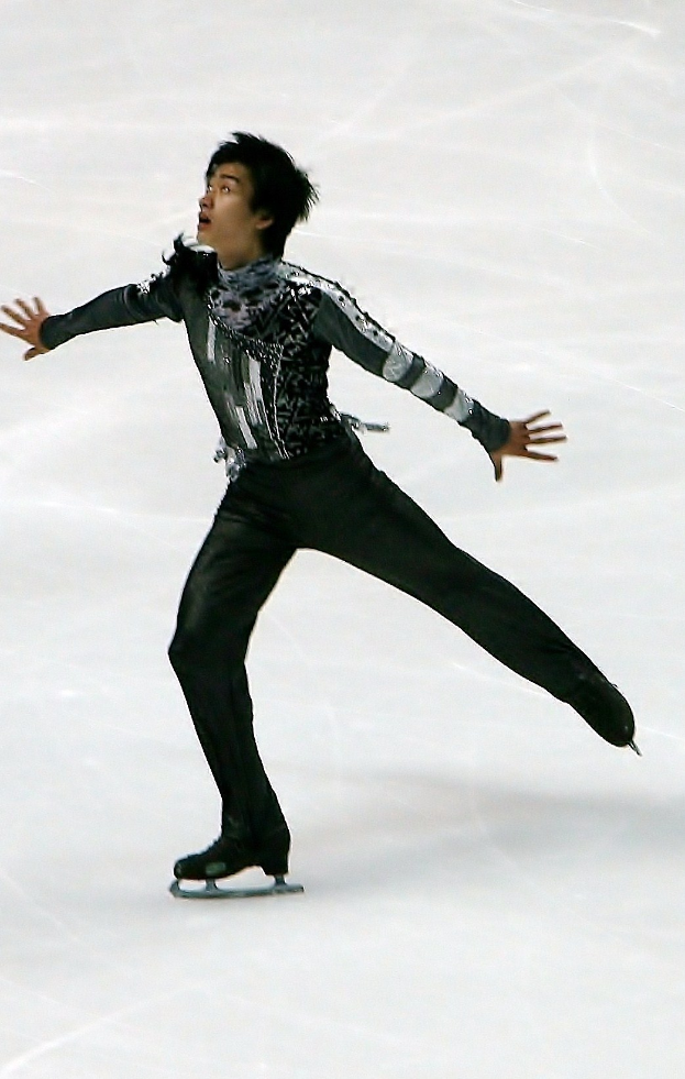 Männlicher Eiskunstläufer in schwarzem Kleid und Eislaufschuhen gleitet über das Eis, führt eine Choreographie mit synchronen Arm- und Beinbewegungen aus.