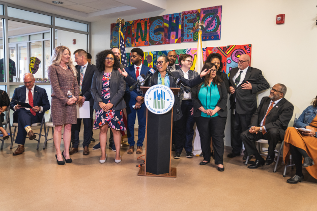 Eine Gruppe von Menschen, die vor einem Rednerpult mit Mikrofon und einer Tafel mit der Aufschrift "Sancho-Stadtrat tritt dem Stadtrat bei" stehen und sitzen, mit Flaggen, Feuerlöscher und Deckenleuchten an der Rückwand.