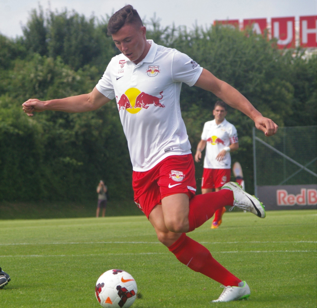 Fußballprofi kickt einen Ball auf einem grünen Rasen mit "RB Leipzig"-Schriftzug im Hintergrund bei klarem blauem Himmel.