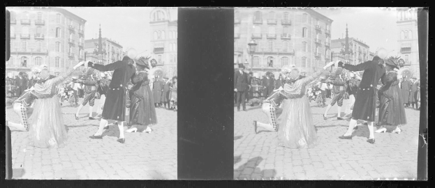 Schwarzes und weißes Foto einer Gruppe von Menschen in Kostümen, die auf der Straße tanzen, umgeben von Gebäuden, Straßenlaternen und Lichtern unter einem klaren Himmel.