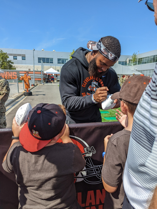 Cleveland Browns Wide Receiver Odell Beckham Jr. unterschreibt Autogramme für Fans auf dem Trainingsgelände des Teams, umgeben von Menschen mit Kappen, einem Banner und Gebäuden im Hintergrund unter einem bewölkten Himmel.