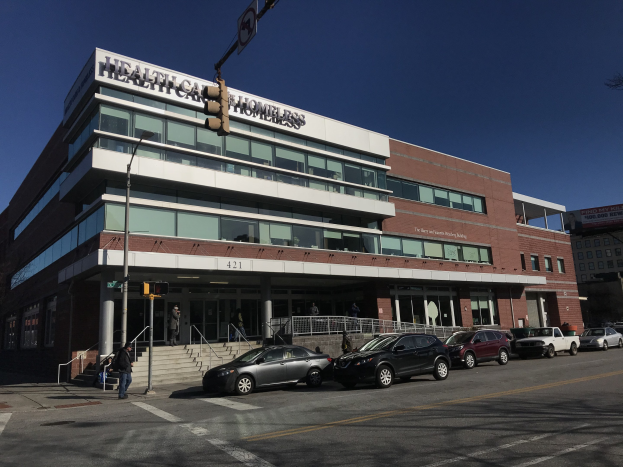 Großes Gebäude als das New York City Health Care Obdachlosenzentrum mit geparkten Autos, Fußgängern, Straßenmöbeln und Bäumen unter einem klaren Himmel.