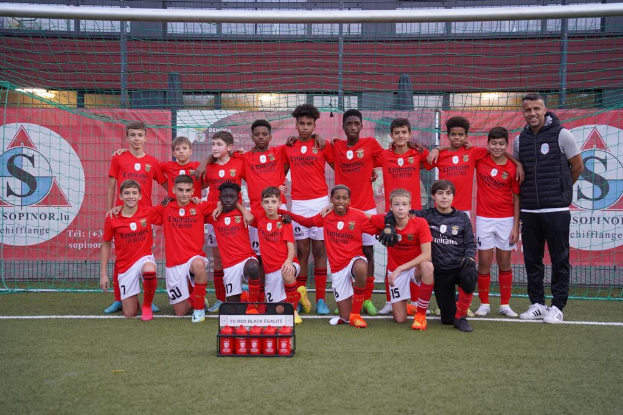 Eine Gruppe junger Männer in roten T-Shirts und schwarzen Shorts steht auf einem Fußballfeld neben einer Kiste roter Gegenstände, im Hintergrund sind Banner mit der Aufschrift "FC Red Black Eglaute - Champions League" zu sehen.