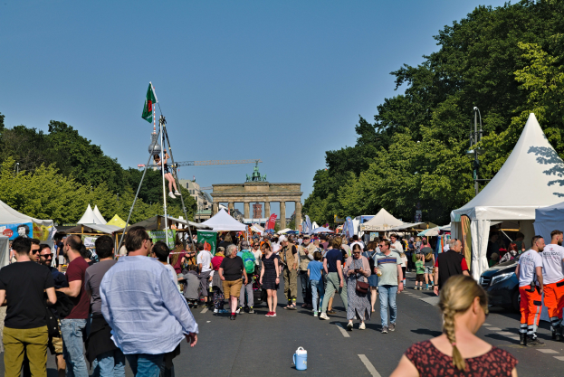 Menschenmenge geht eine Straße mit Zelten, Fahrzeugen und Bäumen entlang bei einem Event mit Fahnenmästern, einem Bogen und einem klaren blauen Himmel.