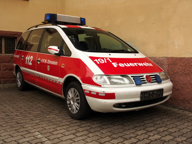 Roter und weißer Minivan mit Text und Zahlen, vor einem Gebäude mit Fenstern geparkt, dient als Ambulanz.
