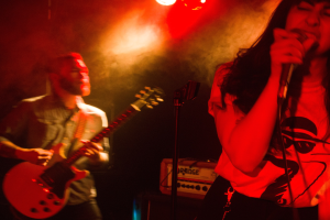 Eine Frau singt in ein Mikrofon auf der Bühne, während ein Mann im Hintergrund eine Gitarre spielt, beleuchtet von Deckenlampen.
