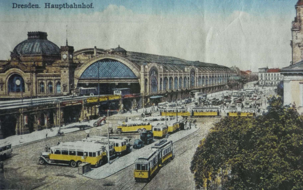 Ein sepiafarbenes Postkartenbild des Dresdner Hauptbahnhof Bahnhofs in Deutschland, das eine belebte Straße mit Fahrzeugen, Fußgängern, Bäumen und Gebäuden unter einem bewölkten Himmel zeigt.