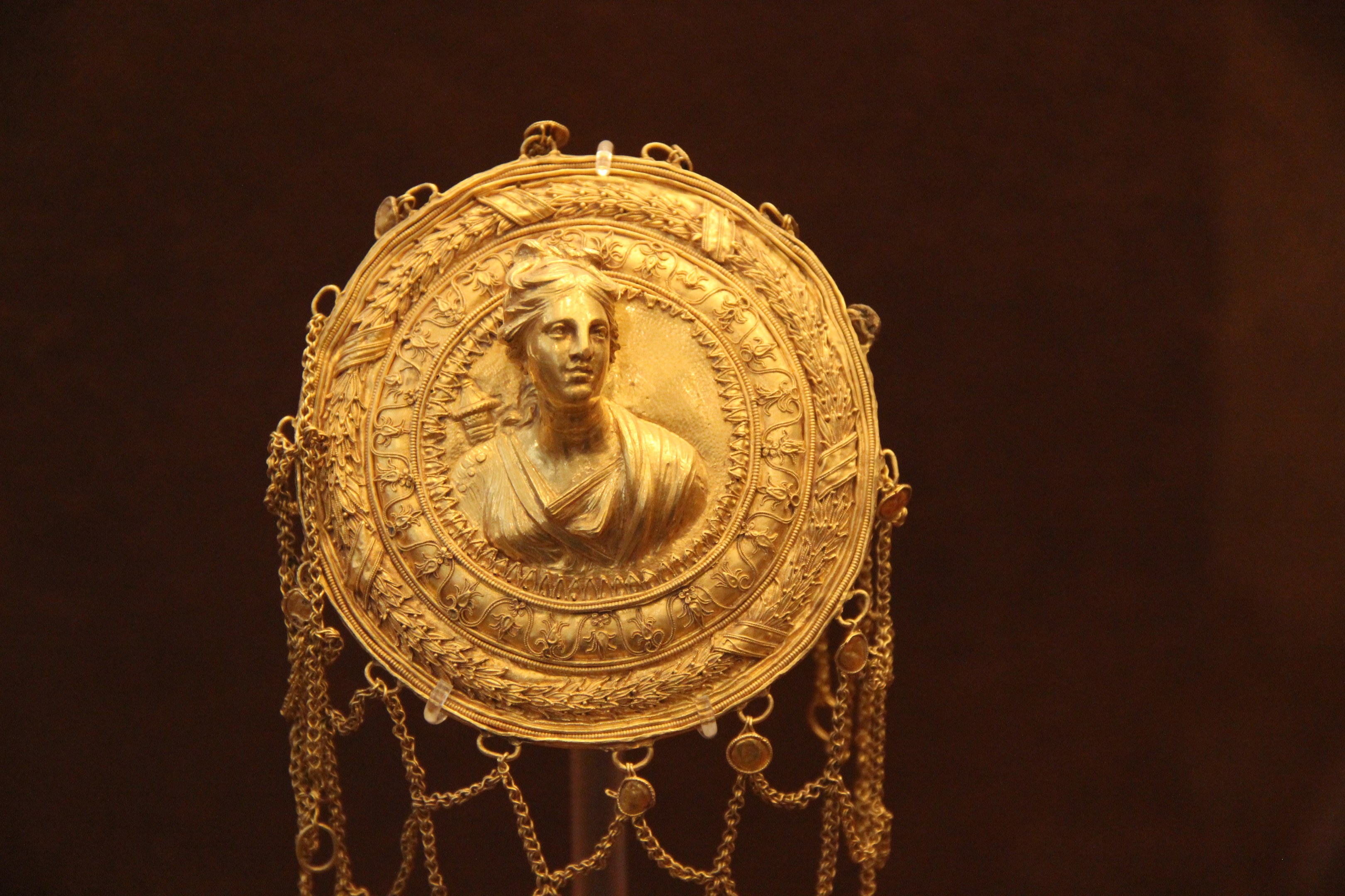 Goldene Medaille mit einer Frauengestalt, die detaillierte Verzierungen zeigt, vor dunklem Hintergrund.