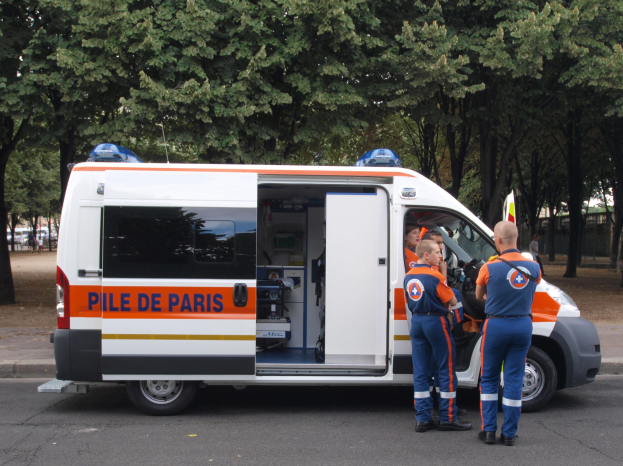 Ambulanz am Straßenrand mit drei Personen davor geparkt, Bäume, Gebäude und andere Fahrzeuge im Hintergrund.