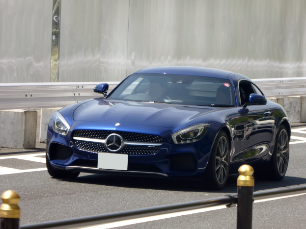 Blauer Mercedes-Benz AMG GT Coupé auf einer Straße fahrend, mit einer Wand im Hintergrund; schlanker Design mit langer Motorhaube, niedriger Karosserie, eckigen Scheinwerfern, großem Kühlergrill, glänzenden Rädern und luxuriösem Lederinnenraum.