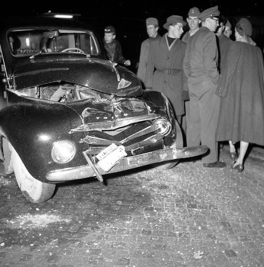 Schwarz-weißes Bild eines stark beschädigten Autos am Straßenrand mit einer Gruppe von Menschen, die es untersuchen.