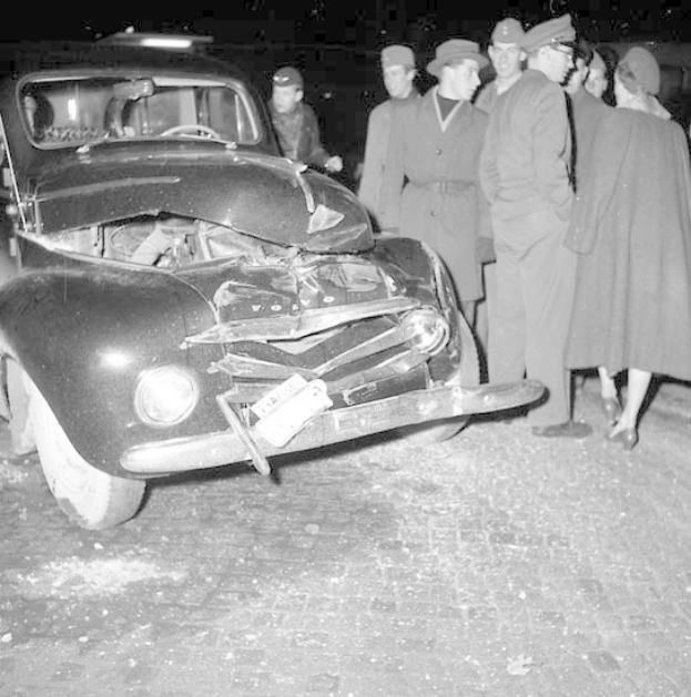 Schwarz-weißes Bild eines stark beschädigten Autos am Straßenrand mit einer Gruppe von Menschen, die es untersuchen.