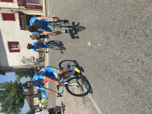 Eine Gruppe von Radfahrern fährt eine Straße entlang an einem Gebäude mit Fenstern und Texttafeln auf der linken Seite vorbei, mit Bäumen und einem klaren Himmel im Hintergrund und Zuschauern, die auf der Straße darunter stehen.
