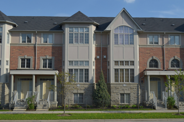 Eine Reihe von Stadthäusern in einem Wohnviertel mit sichtbaren Fenstern, Türen, Säulen, Geländern, Treppen, Pflanzen, Bäumen, Gras, einer Straße im Vordergrund und dem Himmel im Hintergrund.