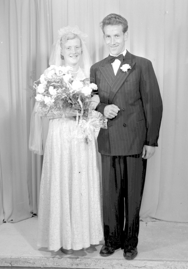 Ein Mann und eine Frau stehen nebeneinander auf einer Bühne, lächeln und die Frau hält einen Blumenstrauß, mit einem Vorhang im Hintergrund.