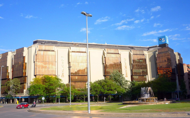 Bancroft-Stadion, ein großes Gebäude mit einem Brunnen davor, umgeben von Straßenlaternen, Straßenlaternen, Kraftfahrzeugen, Fußgängern, Bäumen und einem bewölkten Himmel.