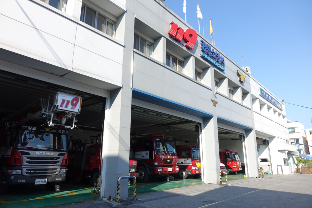 Feuerwache mit mehreren Feuerwehrautos vor der Tür, mit Fenstern, Säulen und Namensschildern, mit Fahnenmasten im Vordergrund, anderen Gebäuden im Hintergrund und einem klaren blauen Himmel.