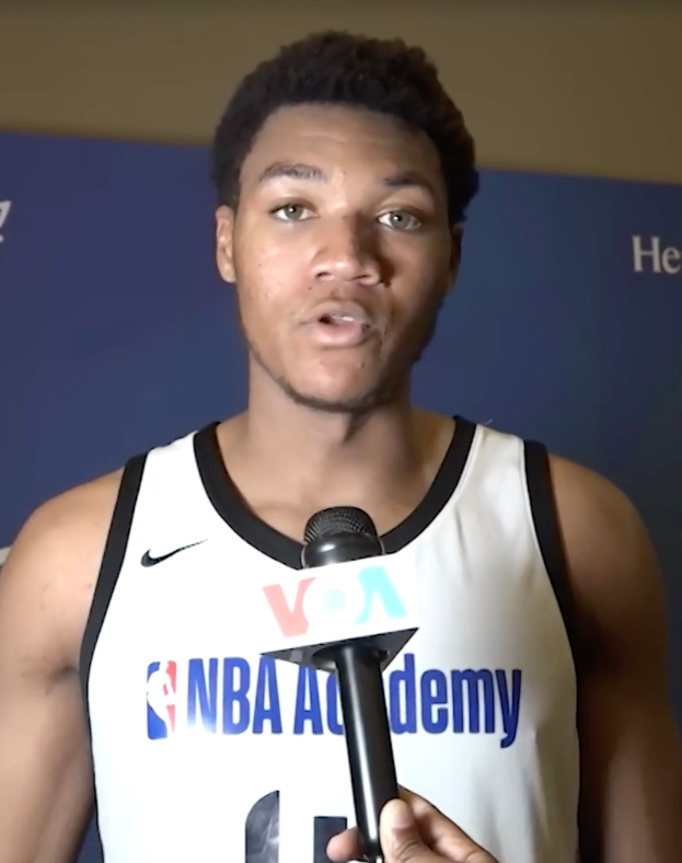 Ein Mann in einem weißen Tanktop hält ein Mikrofon vor ein Banner, auf dem 'NBA All-Star Game 2019' steht und scheint mit der Medien zu sprechen.