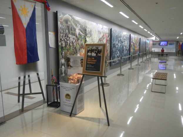 Langer Flur mit einer philippinischen Flagge an der Wand, informative Schautafeln mit Text und Bildern, Ribbon-Ständer, Bänke, Deckenleuchten und ein Bildschirm mit Museums-Informationen im Hintergrund.