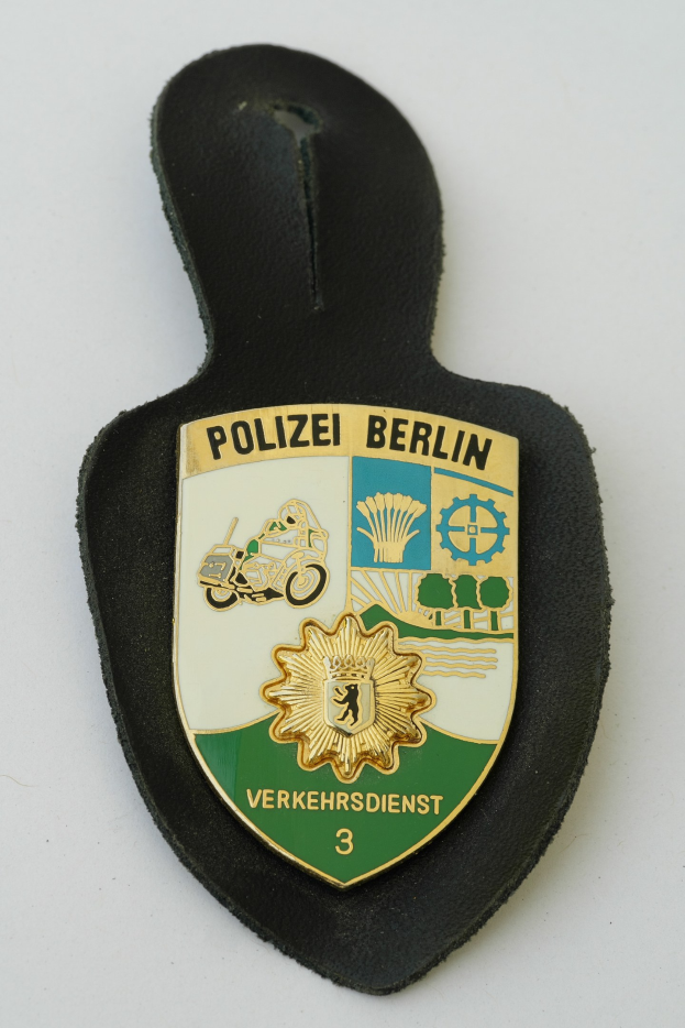 Ein schildförmiges Polizeibadge mit "Polizei Berlin" in schwarzer fettgeschriebener Schrift auf einem blauen Hintergrund mit einem weißen Stern und Rand, das auf einer Oberfläche platziert ist.