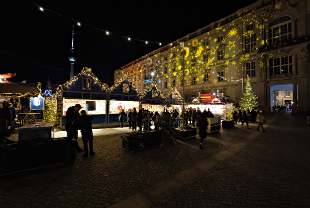 Ein lebendiger Weihnachtsmarkt in Berlin, Deutschland, mit Menschen um hell erleuchtete Stände, festliche Deko und Gebäude mit Fenstern unter einem dunklen Himmel.