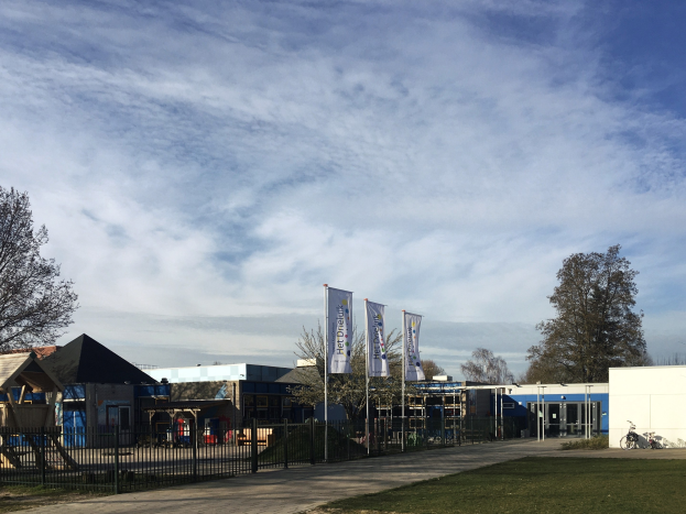 Großes Gebäude mit einem Metallzaun, Rasenfläche, Weg, Bäumen, Pfosten, Bannern, einem Fahrrad, nahen Häusern und einem bewölkten Himmel, identifiziert als der neue Spielplatz an der Schule.