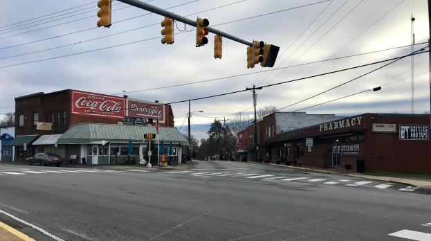 Eine Stadtstra├čenkreuzung mit Ampel, Fahrzeugen, Geb├Ąuden mit Schildern, Strommasten mit Kabeln, B├Ąumen und einem bew├Âlktem Himmel.
