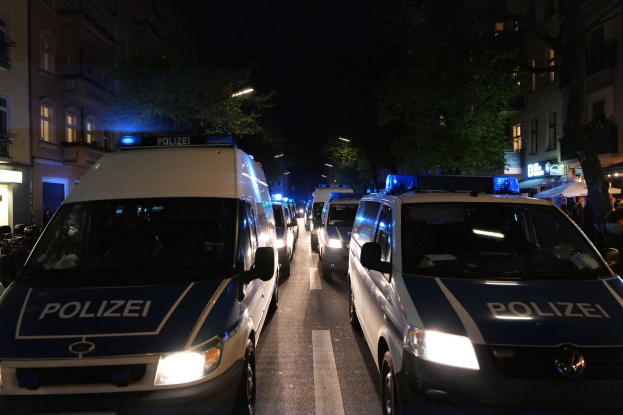 Polizeifahrzeuge parken nachts auf einer Straße mit Text darauf, umgeben von Gebäuden, Bäumen, Laternenmasten und einem dunklen Himmel, mit Menschen, Zelten und anderen Gegenständen auf der rechten Seite.