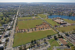Luftbild einer Stadt mit Gebäuden, Bäumen, Gras, Straßen mit Fahrzeugen, Wasser und Wolken, das einen großen zentralen Platz zeigt, der für einen Park vorgesehen ist.