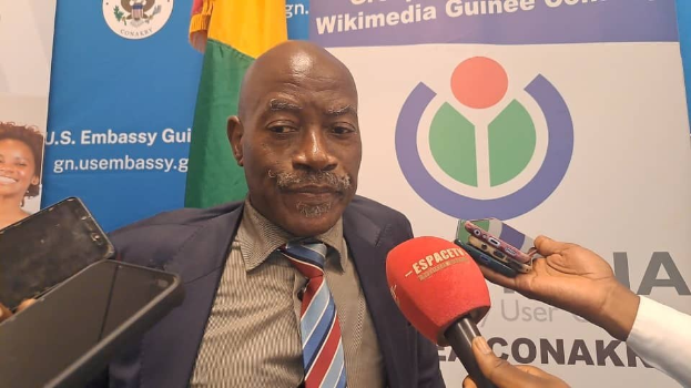 Ein Mann im Anzug spricht mit Reportern, Mikrofon und Gerät im Vordergrund, Banner, Flaggen und eine Frau im Hintergrund.