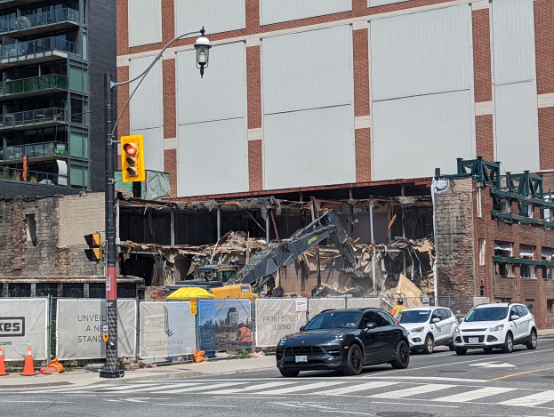 Ein großes Gebäude wird in der Mitte einer Straße abgerissen, mit Fahrzeugen, Verkehrskegeln, einer Ampel und einem Kran im Vordergrund und Gebäuden mit Fenstern im Hintergrund.