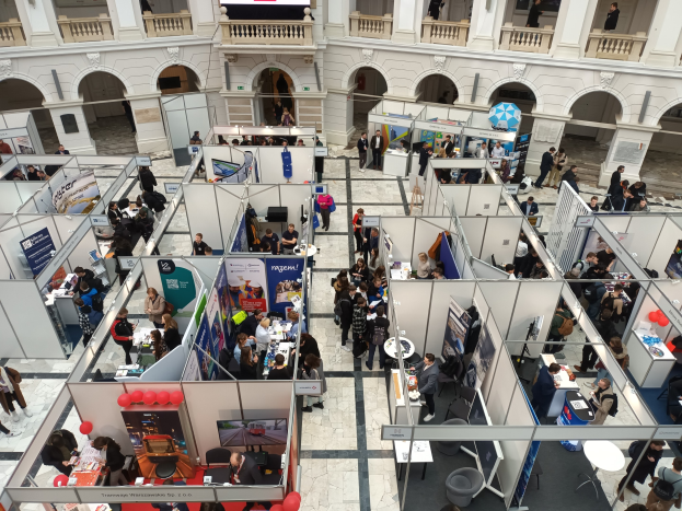 Große Gruppe von Teilnehmern auf einer internationalen Fachmesse mit Ständen, Bannern, Tischen, Stühlen und Luftballons in einem Raum mit architektonischen Bögen und Säulen.