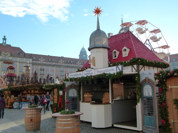 Ein geschäftiger Weihnachtsmarkt in Nürnberg, Deutschland mit Menschen um geschmückte Stände, festlicher Beleuchtung, Schmuck, Gebäuden mit Fenstern, einem Riesenrad, Wolken am Himmel und einer Tafel mit Schrift auf der rechten Seite.