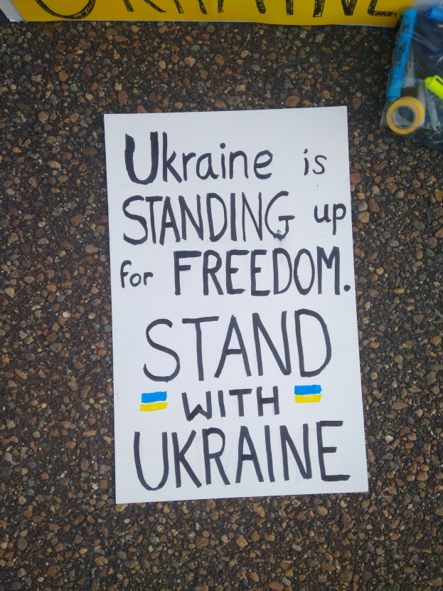 Weißer Zettel mit dem Text "Ukraine steht für Freiheit, steht mit Ukraine" auf dem Boden vor einer gelben Fahne, rechts ein Plastikbehälter mit verschiedenen Gegenständen.