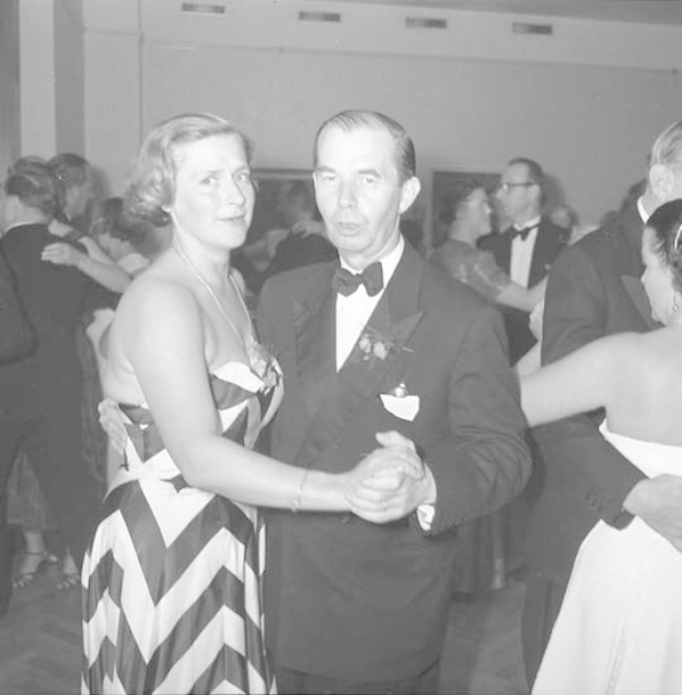 Zwei Menschen, ein Mann in einem schwarzen Blazer und weißem Hemd mit schwarzer Hose und eine Frau in einem unbekannten Outfit, tanzen zusammen auf einer formellen Veranstaltung, indem sie sich an den Händen halten und zur Musik wiegen.