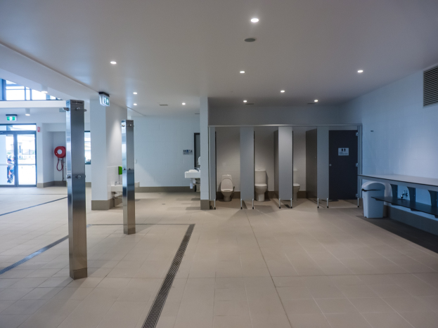 Große Öffentliche Toilette mit mehreren Toiletten, tragenden Säulen, einem Mülleimer, einer Tür, einem Schild, einem Feuerlöscher und deckenmontierten Leuchten.