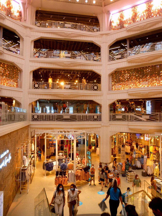 Ein belebtes Einkaufszentrum mit Menschen, die durch eine Halle gehen, Schaufensterpuppen mit Kleidern, Geländer, Lampen und Texttafeln an den Wänden.