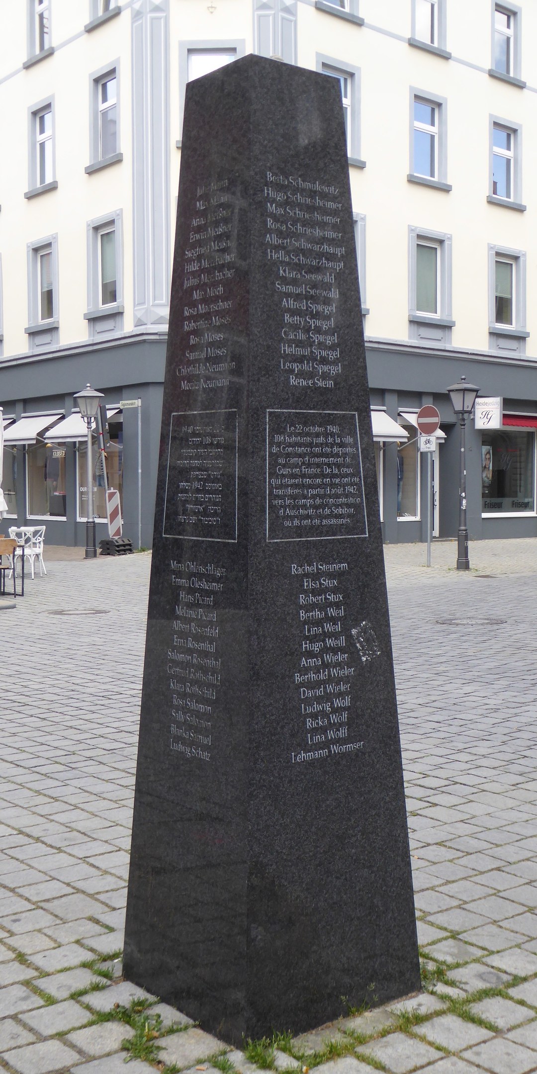 Ein hohes Denkmal mit eingravierter Inschrift steht auf einem Stadtplatz, umgeben von Gebäuden, Straßenmöbeln und Menschen unter einem sichtbaren Himmel.