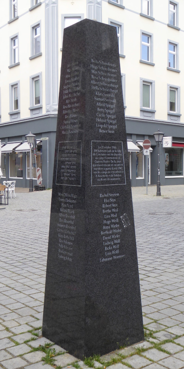 Ein hohes Denkmal mit eingravierter Inschrift steht auf einem Stadtplatz, umgeben von Gebäuden, Straßenmöbeln und Menschen unter einem sichtbaren Himmel.