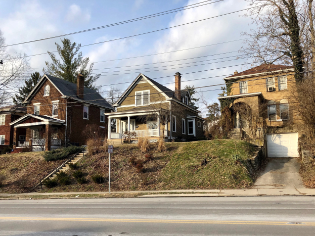 Reihe von Häusern entlang einer Straße mit Fenstern, Türen, Säulen, Geländern, Stufen, Pflanzen, Gras, Bäumen und einem Pfahl mit einer Tafel.