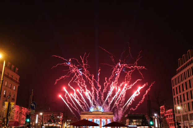 Eine belebte Stadtstraße während einer Silvesterfeier in Berlin, mit Menschen, Fahrzeugen, Gebäuden und Feuerwerk, das den Nachthimmel erhellt.