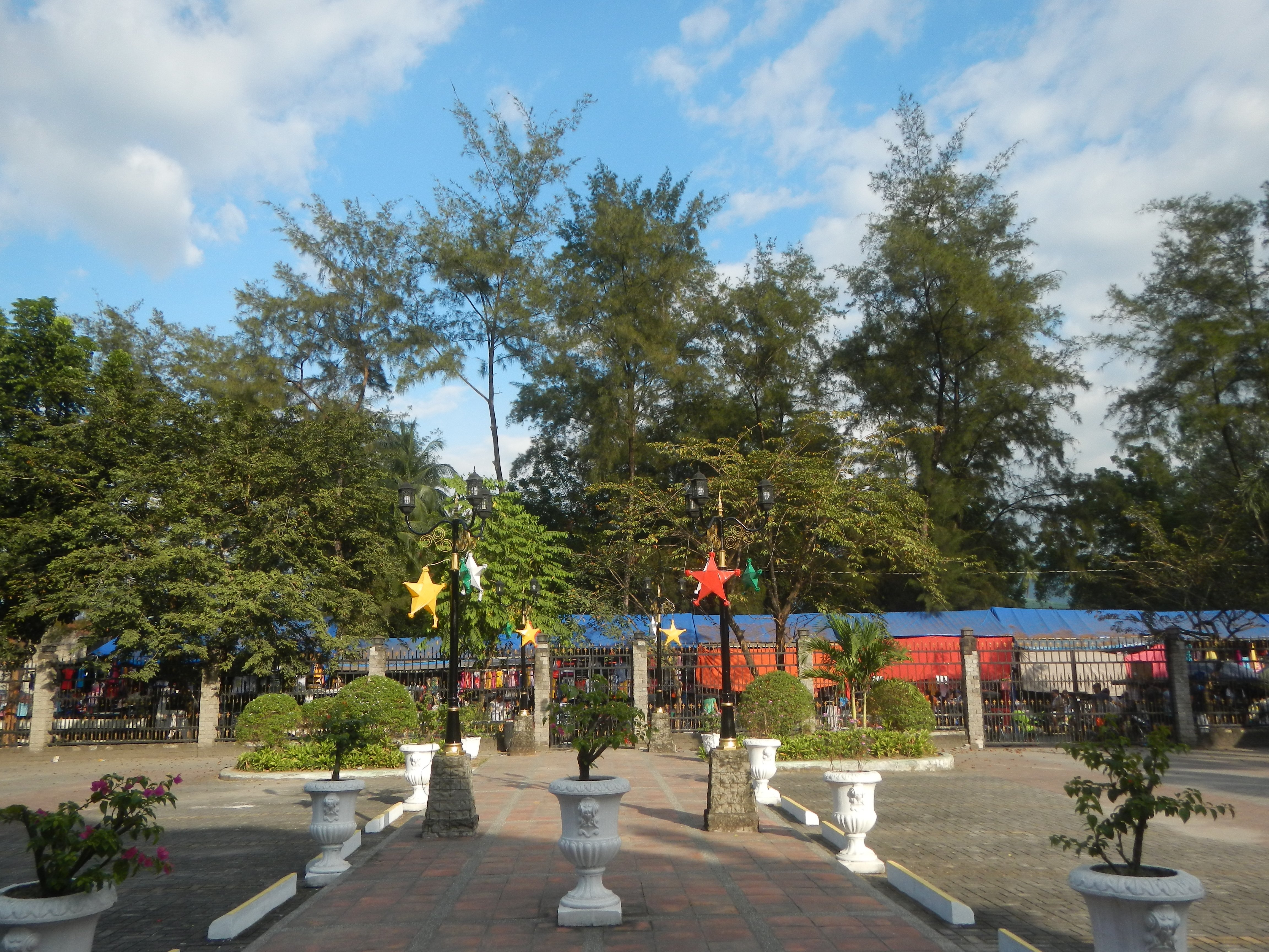 Ein Park mit Topfpflanzen und Bäumen, umgeben von Straßenlaternen, Dekorationen und Grills, mit einem bewölkten Himmel im Hintergrund.