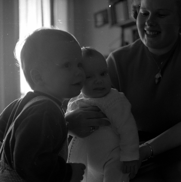 Eine Frau hält ein Baby mit einem warmen Lächeln, mit zwei anderen Kindern, die daneben stehen, in einem gemütlichen Raum mit Rahmen und einem Fenster im Hintergrund.